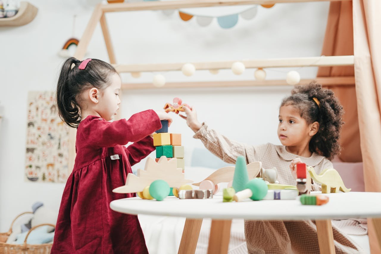 services-07 Two young girls enjoying playtime with wooden toys indoors in a warm, colorful playroom.