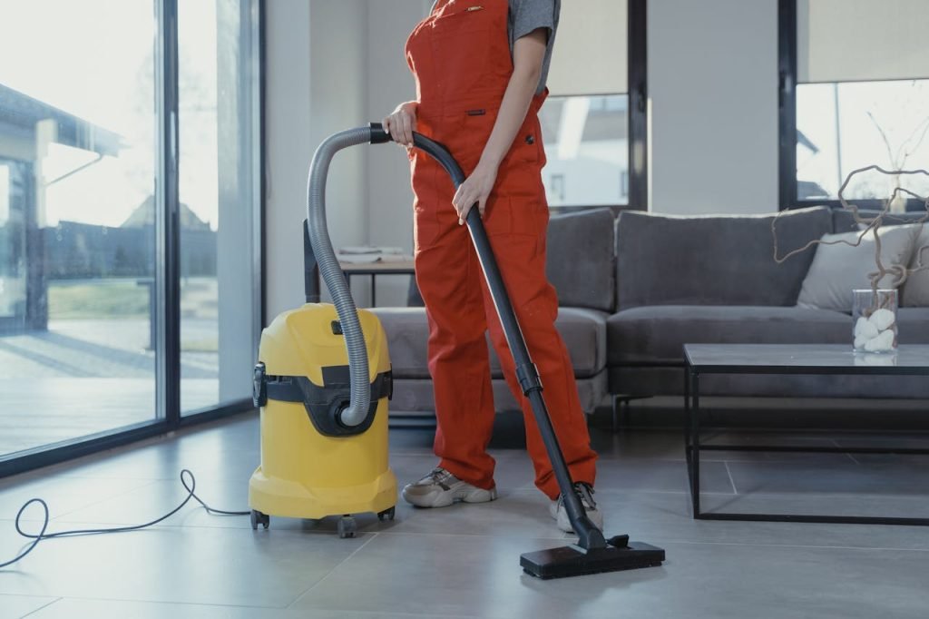 person-vacuuming-the-floor-6195275 A professional cleaner in red coveralls vacuuming a modern living room with a yellow vacuum cleaner.